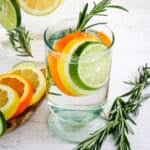 A glass of rosemary citrus water with fresh rosemary sprigs on the side and sliced lemon, lime and orange in the glass.
