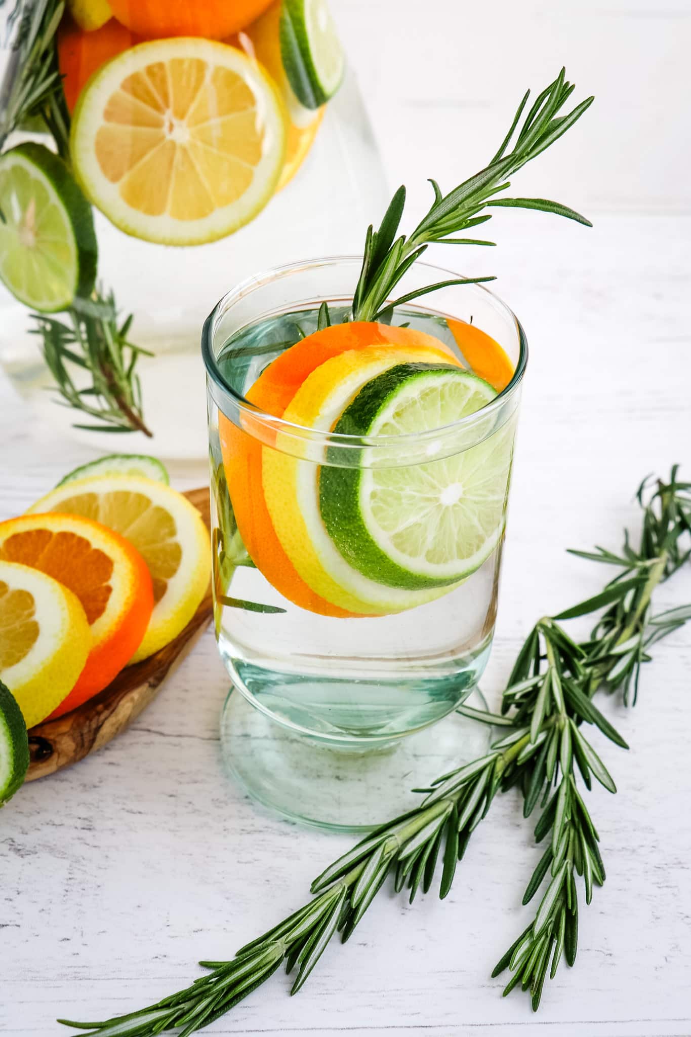 A glass of rosemary citrus water with fresh rosemary sprigs on the side and sliced lemon, lime and orange in the glass.