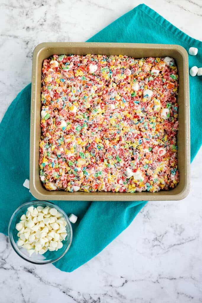 Fruity pebbles marshmallow treats pressed into a square pan with white chocolate chips on the side.