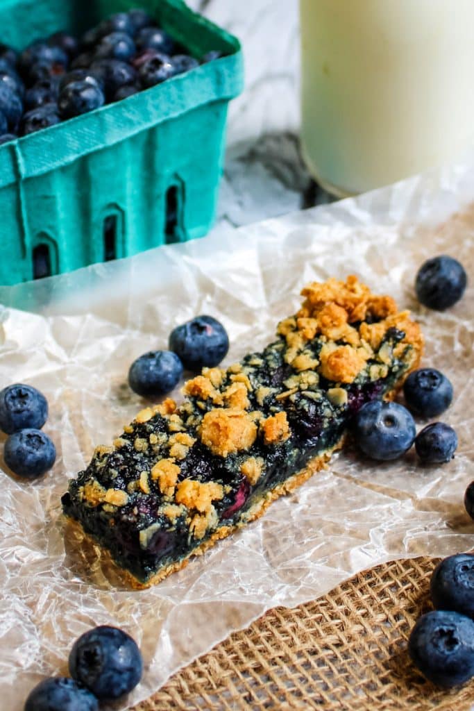 A single blueberry oatmeal bar with fresh blueberries on the side, with a small bottle of milk in the background.