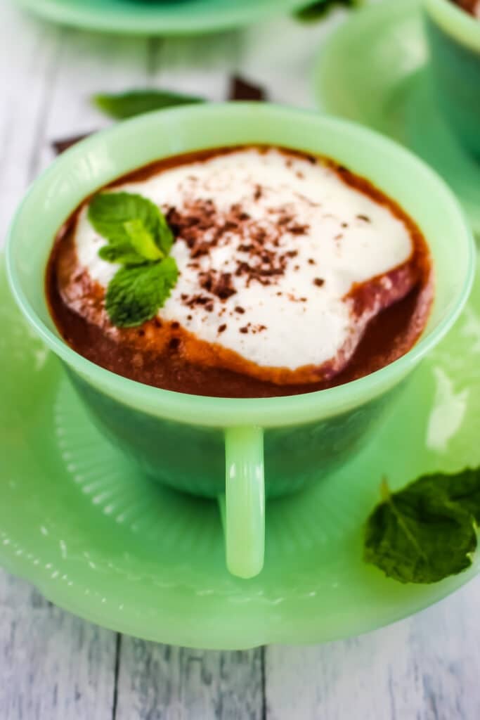 Cup of mint hot chocolate in green cup and saucer, topped with whipped cream and mint leaves.