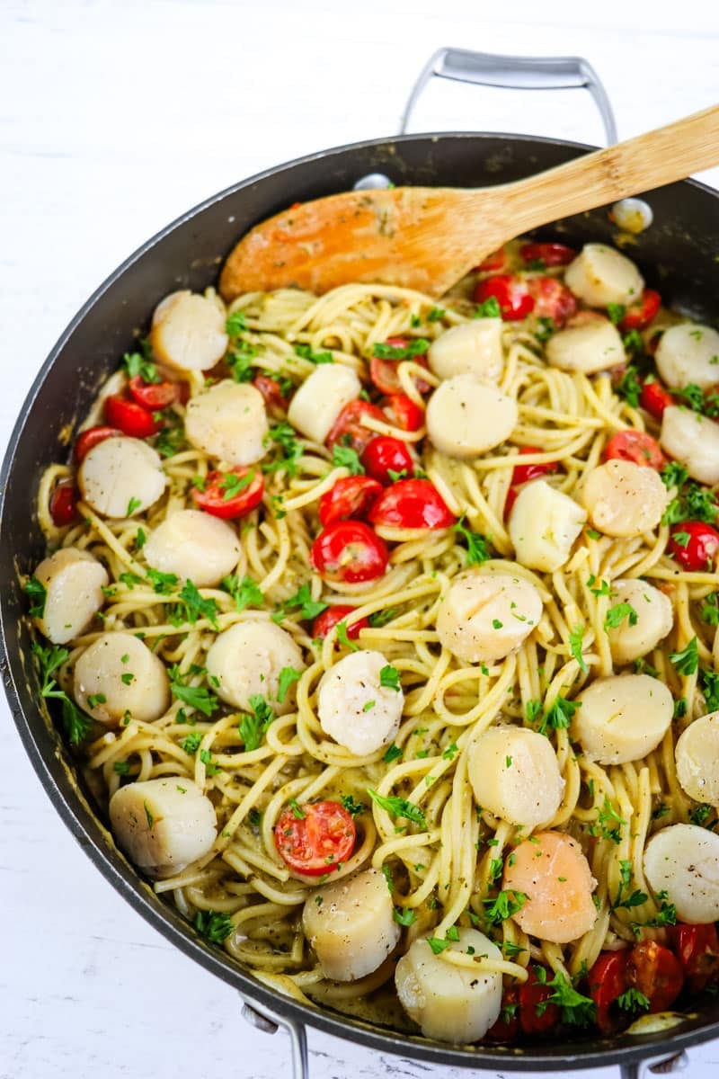 Creamy pesto pasta with scallops in a large skillet with wooden spoon on the side.