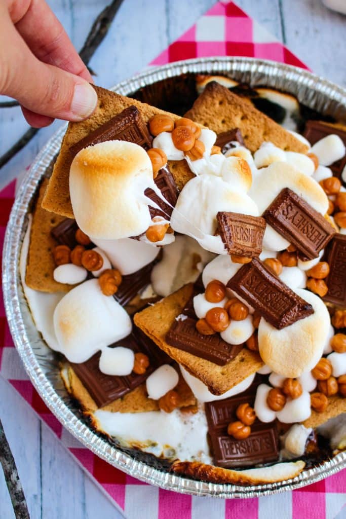 Campfire smores nachos made in a round pan with single cracker being pulled from the pan with marshmallow and chocolate on top.
