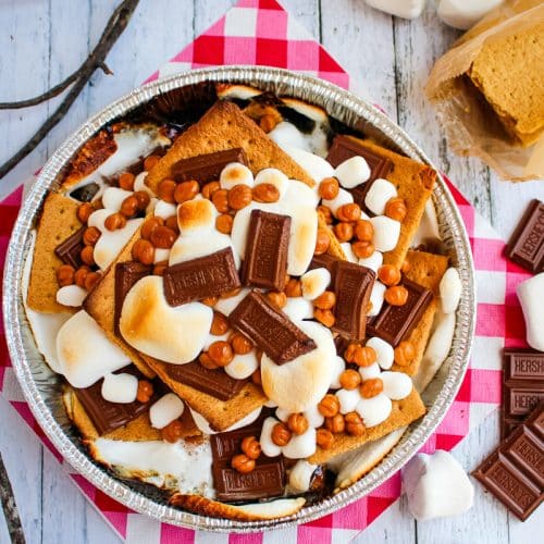 Campfire smores in pan topped with caramel bits, and sticks with marshmallows on the side.
