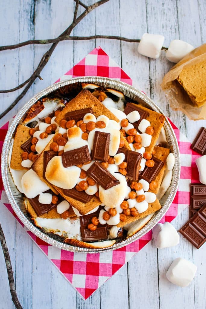 Campfire smores in pan topped with caramel bits, and sticks with marshmallows on the side.