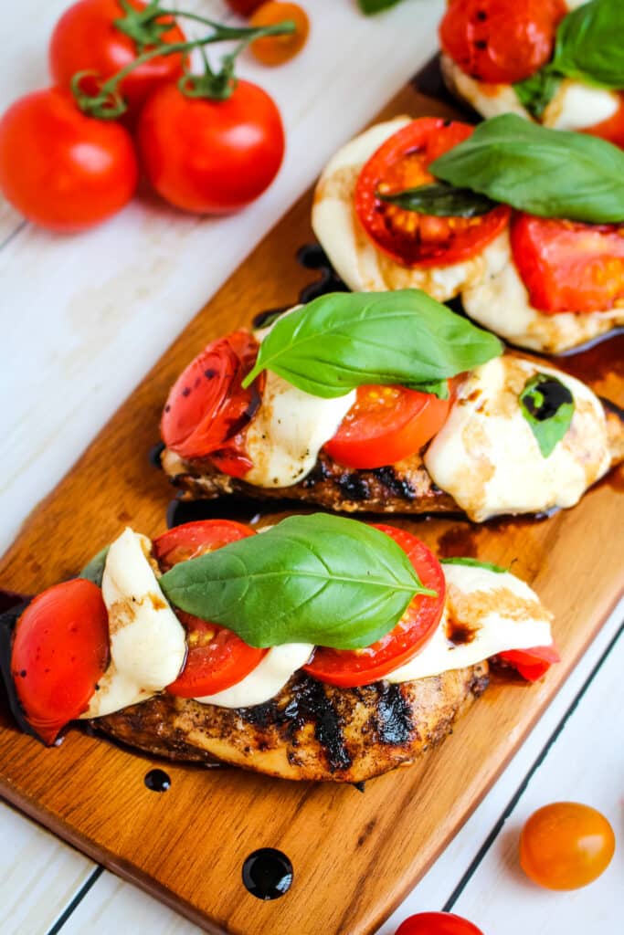 Grilled caprese chicken on a cutting board, topped with fresh basil leaves and tomatoes on the side.
