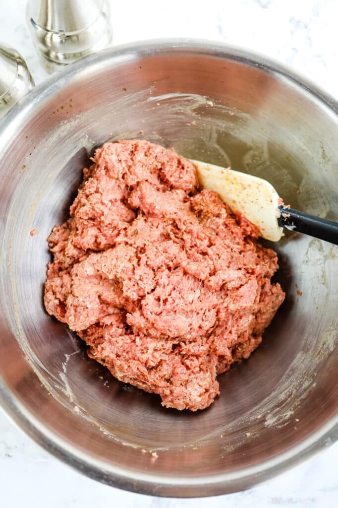 Ground beef mixture in a mixing bowl, with a rubber scraper.