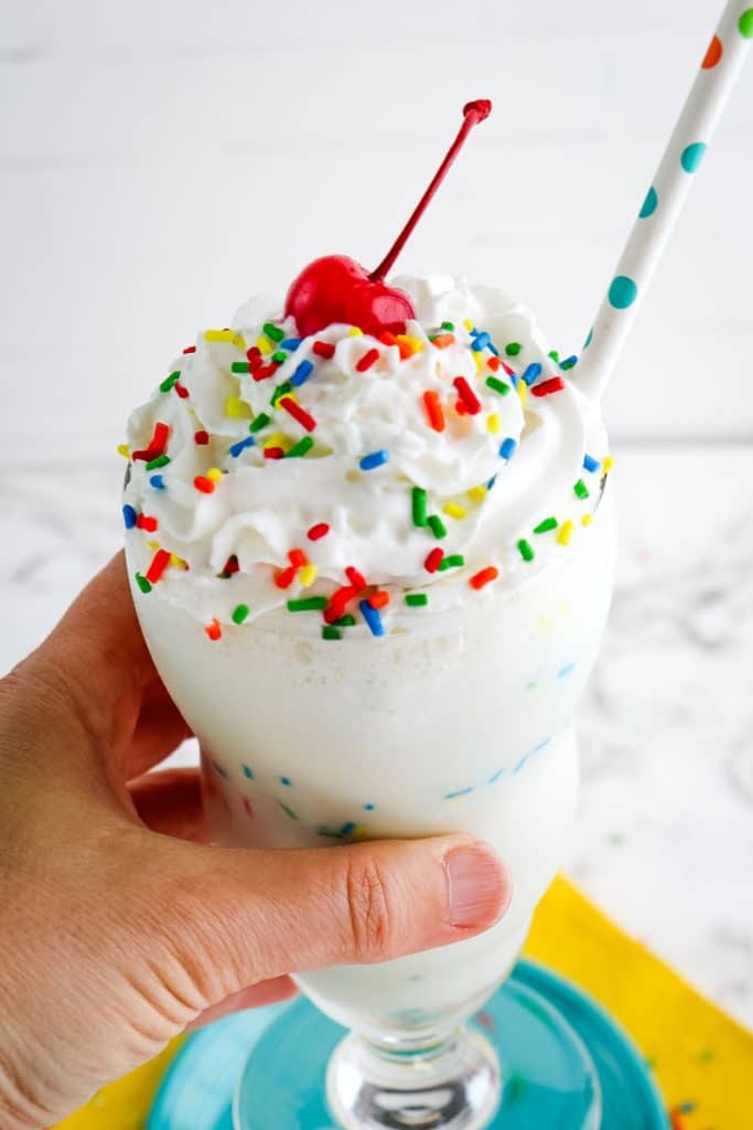 A birthday cake milkshake held in hand and topped with whipped cream, sprinkles and a maraschino cherry.