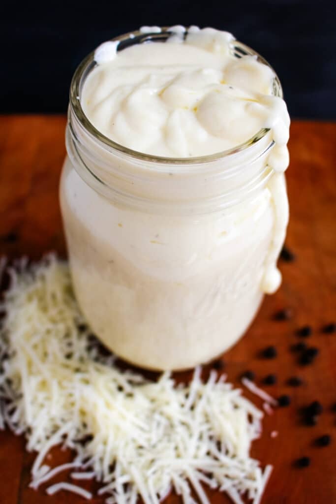Homemade Alfredo sauce in a jar with shredded cheese and peppercorns on the side.