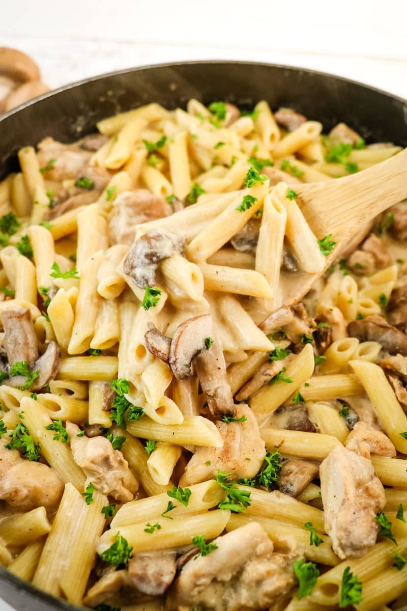 Creamy chicken and mushroom pasta being spooned out of the skillet with a wooden spoon.