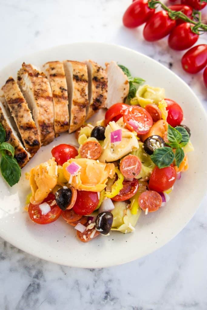 Italian tortellini pasta salad on a plate with grilled chicken and fresh basil leaves for garnish.