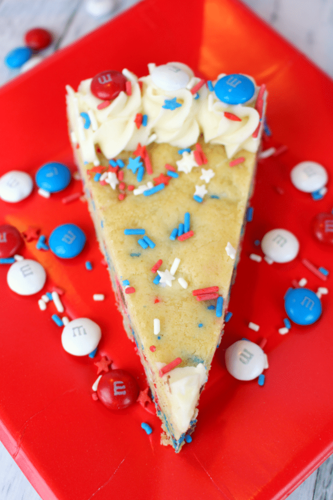 close up photo of Slice of red white and blue sugar cookie cake on red plate with extra M&M's and sprinkles.