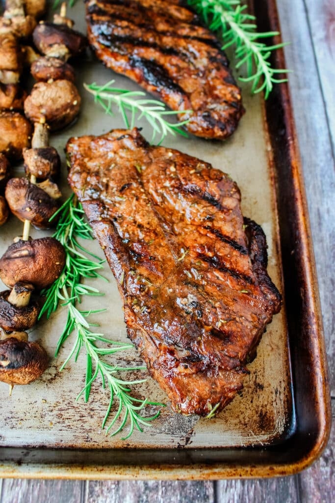 Grilled steak on a pan with fresh rosemary sprigs and grilled mushrooms on the side.