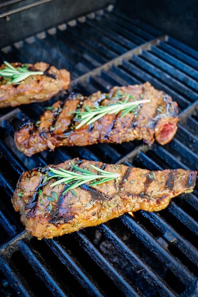 Steaks on a grill topped with fresh rosemary sprigs.