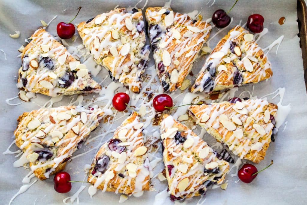 Cherry almond scones on a parchment paper lined pan, glazed and garnished with fresh cherries.