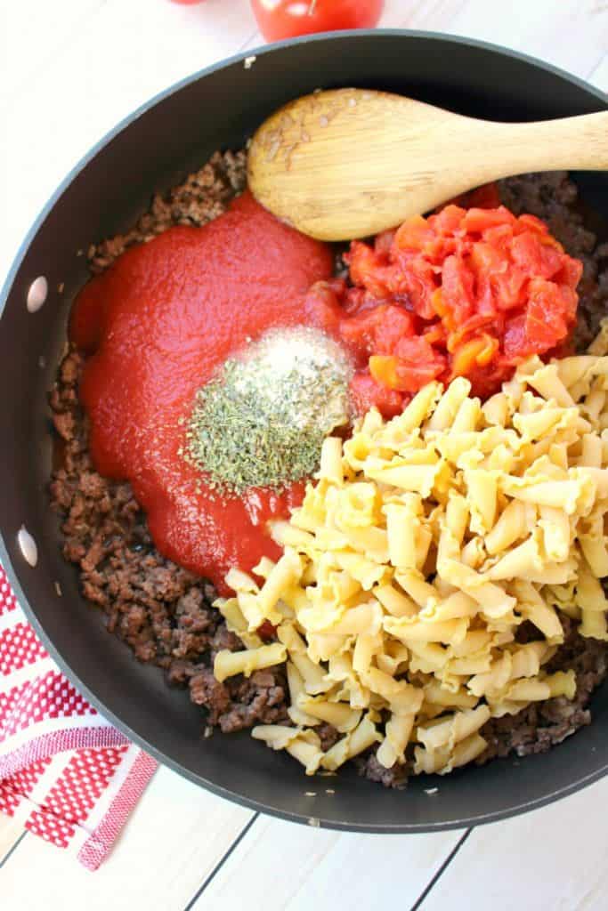 Ground beef in skillet with added ingredients of tomato sauce, diced tomatoes, uncooked pasta and spices.