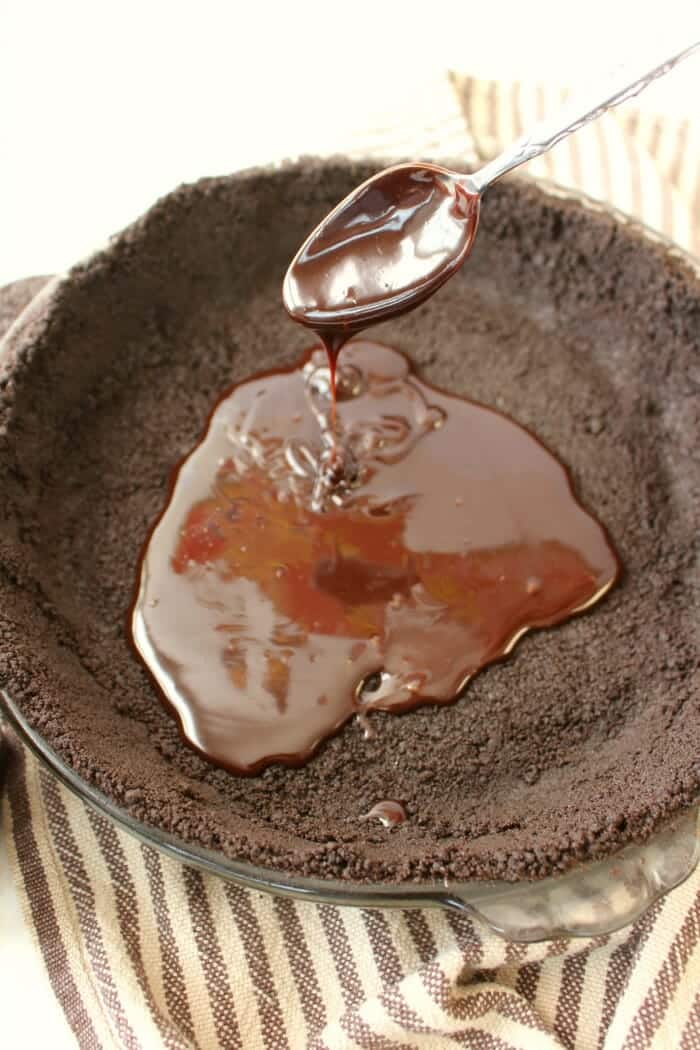 Hot fudge ice cream topping being spooned into oreo cookie crust in pie plate.