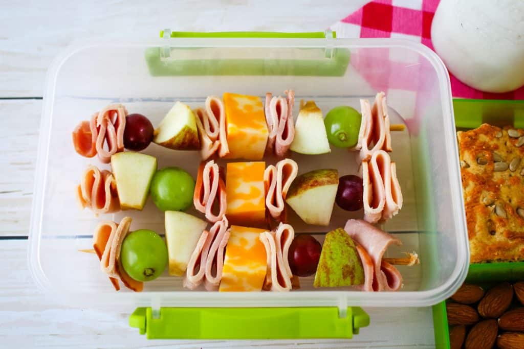 Meat and cheese kabobs in a lunchbox container with crackers and milk on the side.