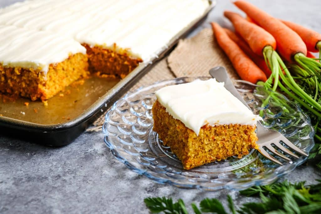 Slice of carrot cake on a glass plate, with whole cake in the background.