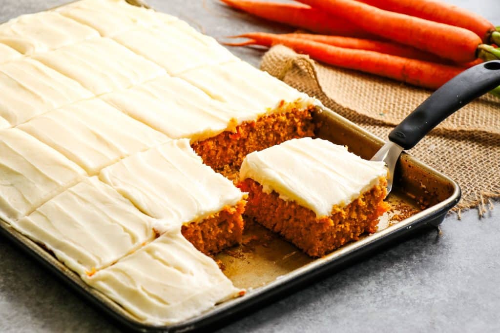 Carrot cake sheet cake, cut into slices in the pan, with one slice on a serving utensil.