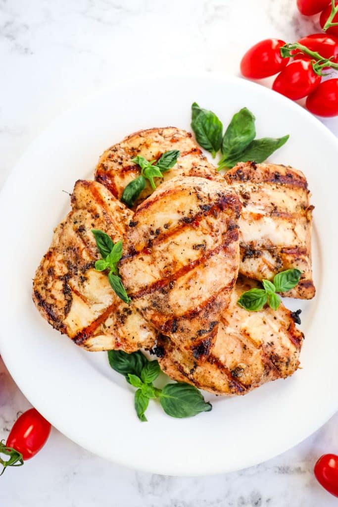 Italian grilled chicken breasts on a white plate garnished with fresh basil leaves and cherry tomatoes on the side.