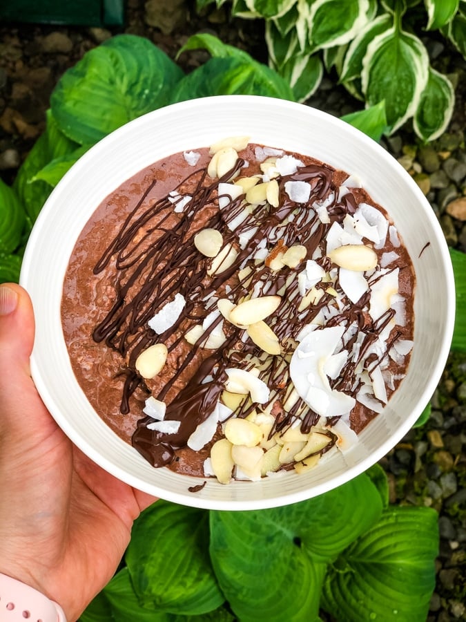 Chocolate protein oats in a white bowl topped with coconut, almonds and chocolate drizzle.