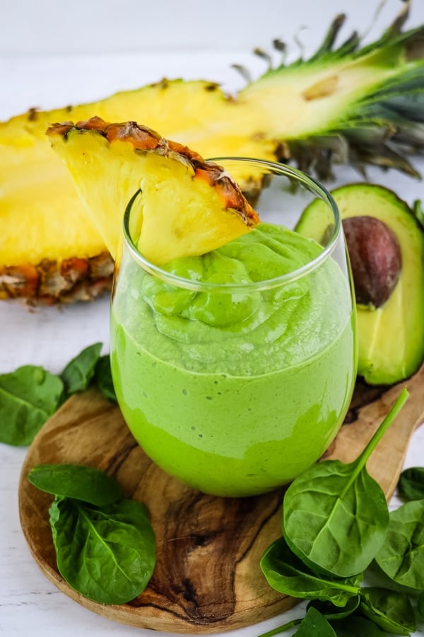 Green superfood smoothie in glass with pineapple slice garnish and spinach leaves on the side.