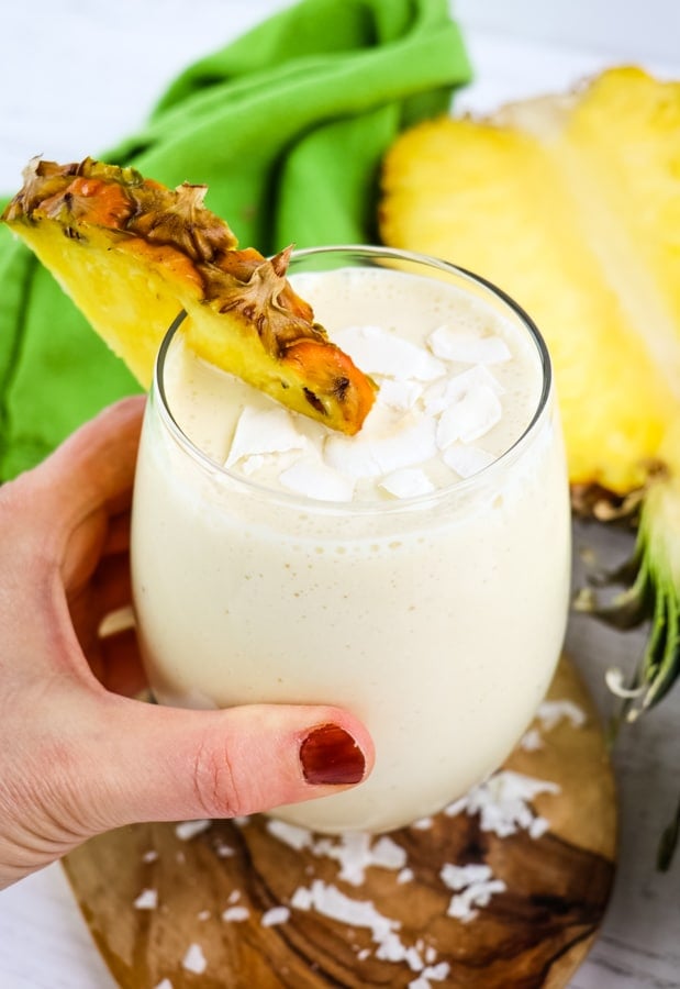 Pineapple coconut smoothie garnished with pineapple slice and coconut flakes held in hand.