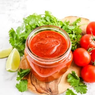 Homemade taco sauce in a short mason jar without lid. Tomatoes, cilantro and lime wedges for garnish.