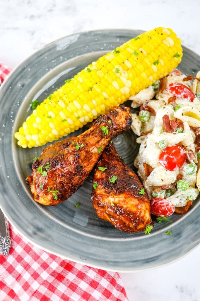 BBQ drumsticks on plate with corn on the cob and pasta salad.