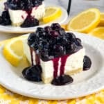 Slice of lemon blueberry cheesecake on plate, topped with blueberry topping, garnished with lemons.