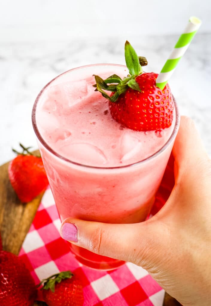 Pink drink with straw and strawberry garnish, being held in hand.