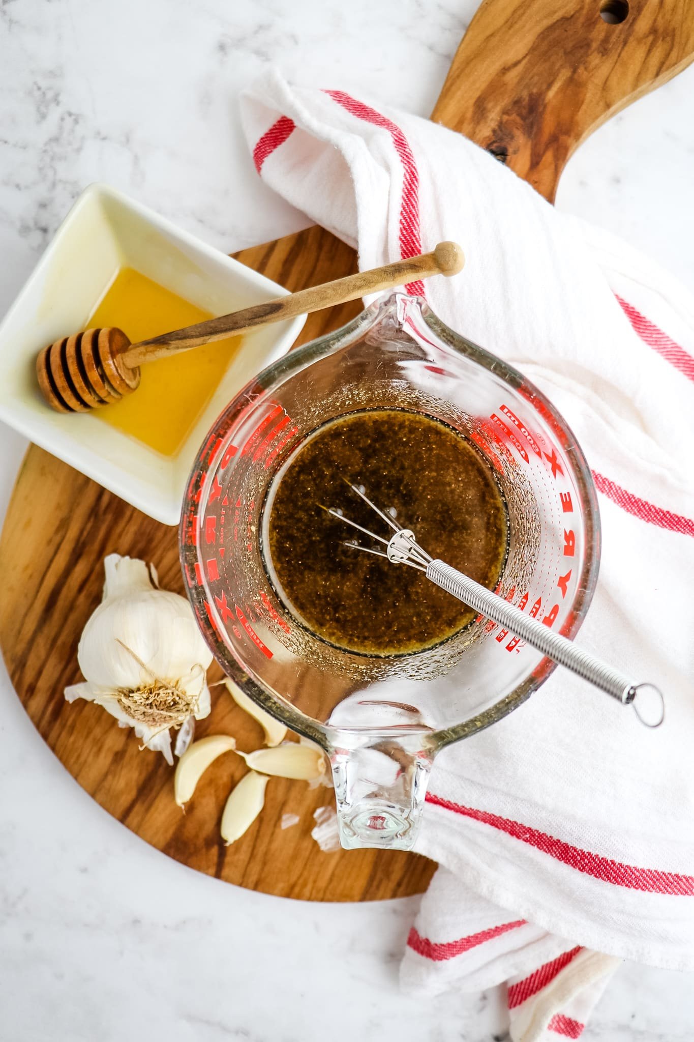 Honey garlic marinade in measuring cup with small whisk, garlic cloves and honey on the side.