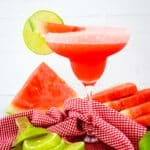 Frozen watermelon margarita, in margarita glass with limes and watermelon on the side.