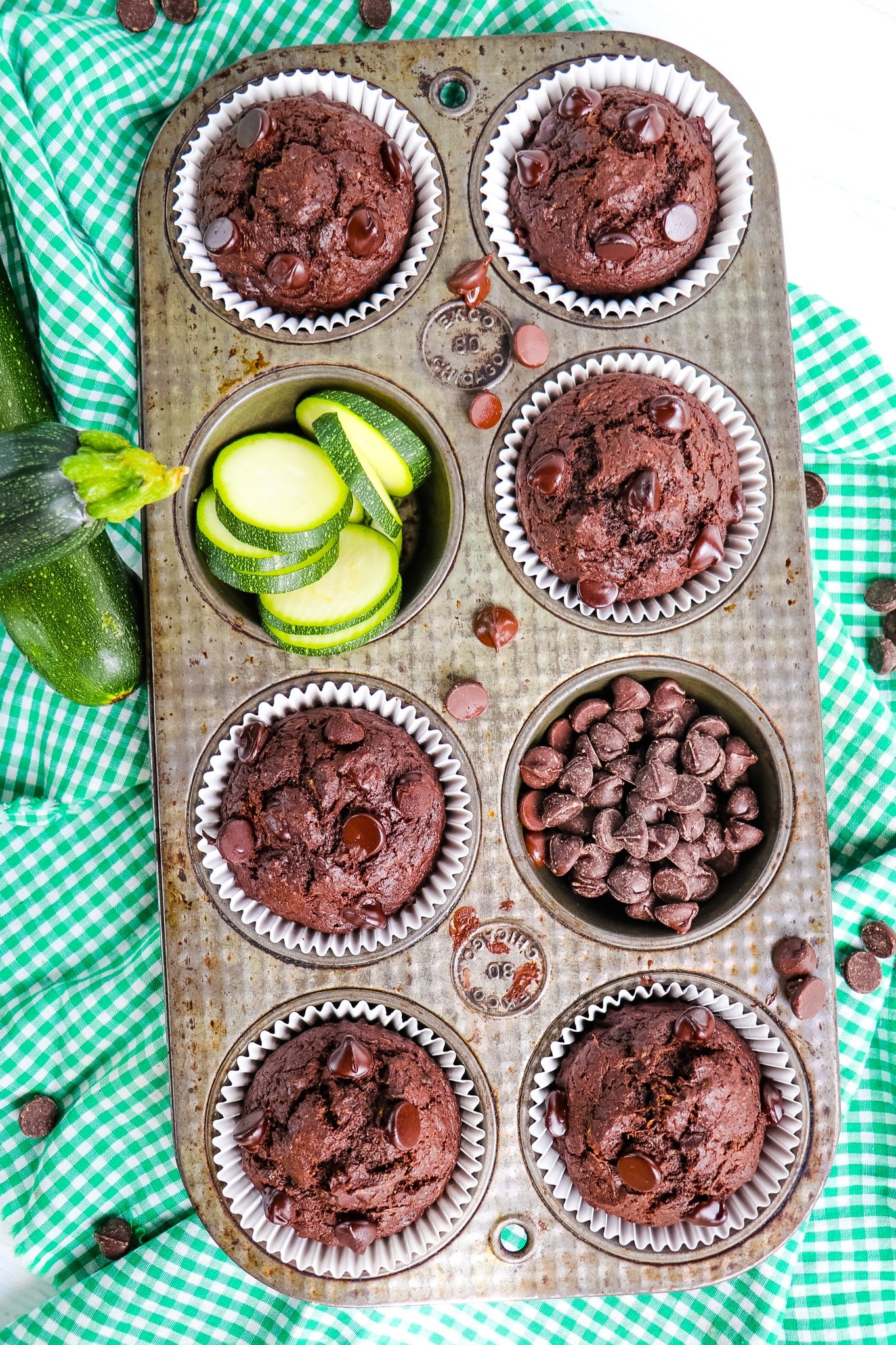 Chocolate zucchini muffins in muffin tin with chocolate chips and sliced zucchini.