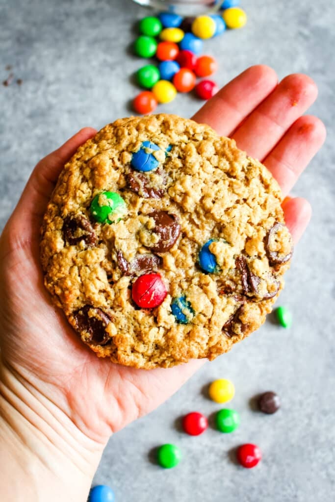 Large monster cookie held in hand, with M&M candies in the background.