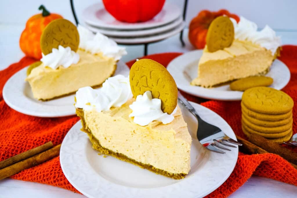 Three slices of no bake pumpkin pie cheesecake on plates, garnished with whipped cream and ginger snap cookies.
