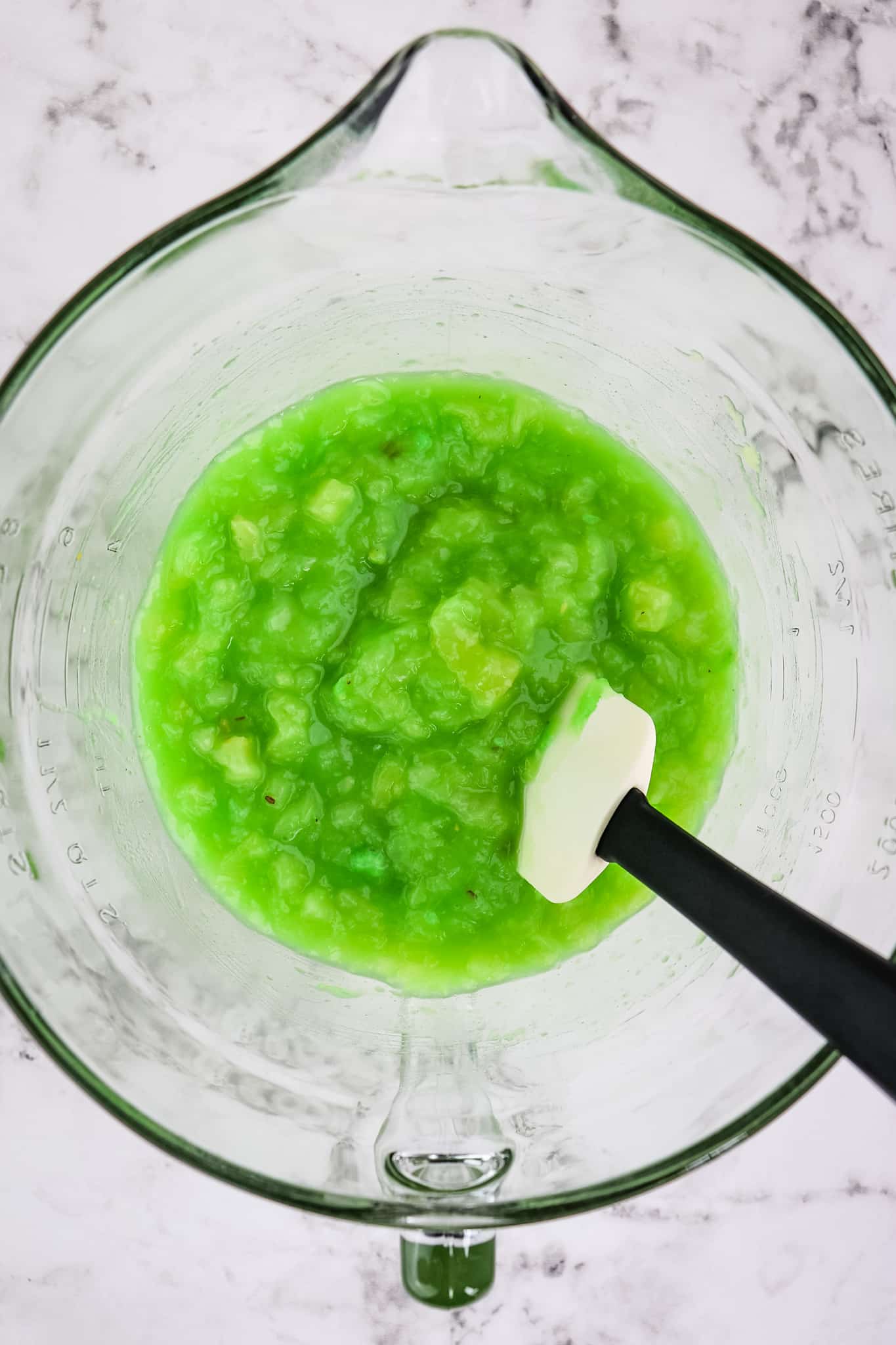 Pistachio pudding mixed together with crushed pineapple in a mixing bowl with rubber scraper on the side.