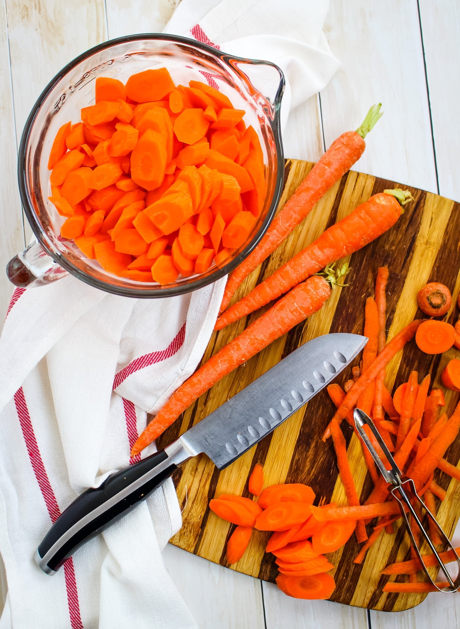 Whole carrots being peeled and sliced on a cutting board. Bowl of sliced carrots on the side.