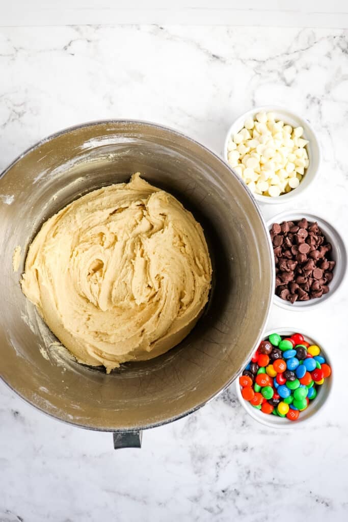 Cookie dough in a mixing bowl with M&M's, chocolate chips and white chocolate chips on the side.