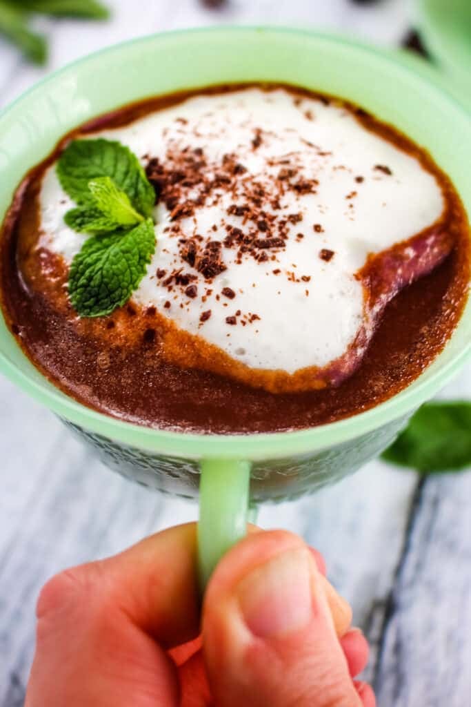 Cup of mint hot chocolate held in hand with a sprig of mint and whipped cream.