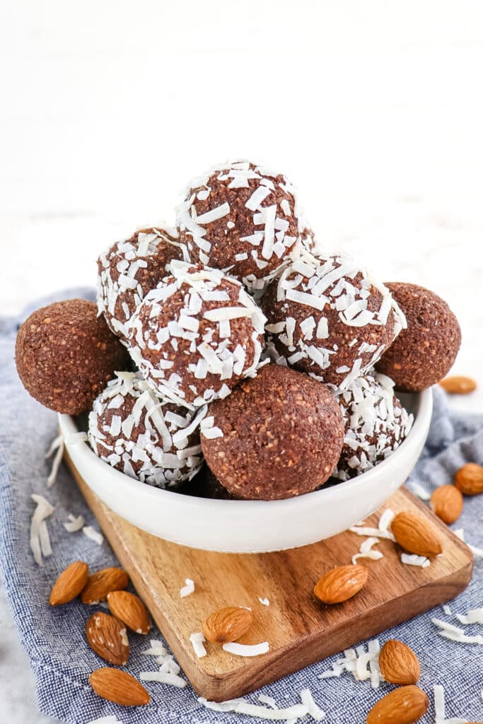 Bowl of chocolate and coconut date balls with almonds and coconut garnishes on the side.