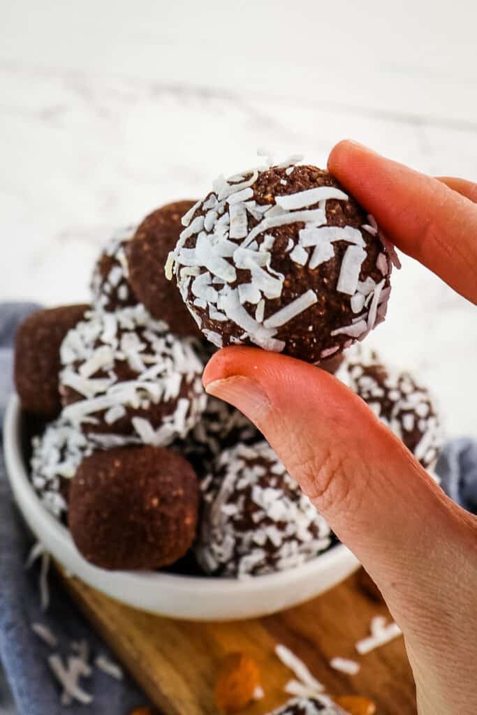 Coconut date ball held in hand with bowl of date balls in the background.