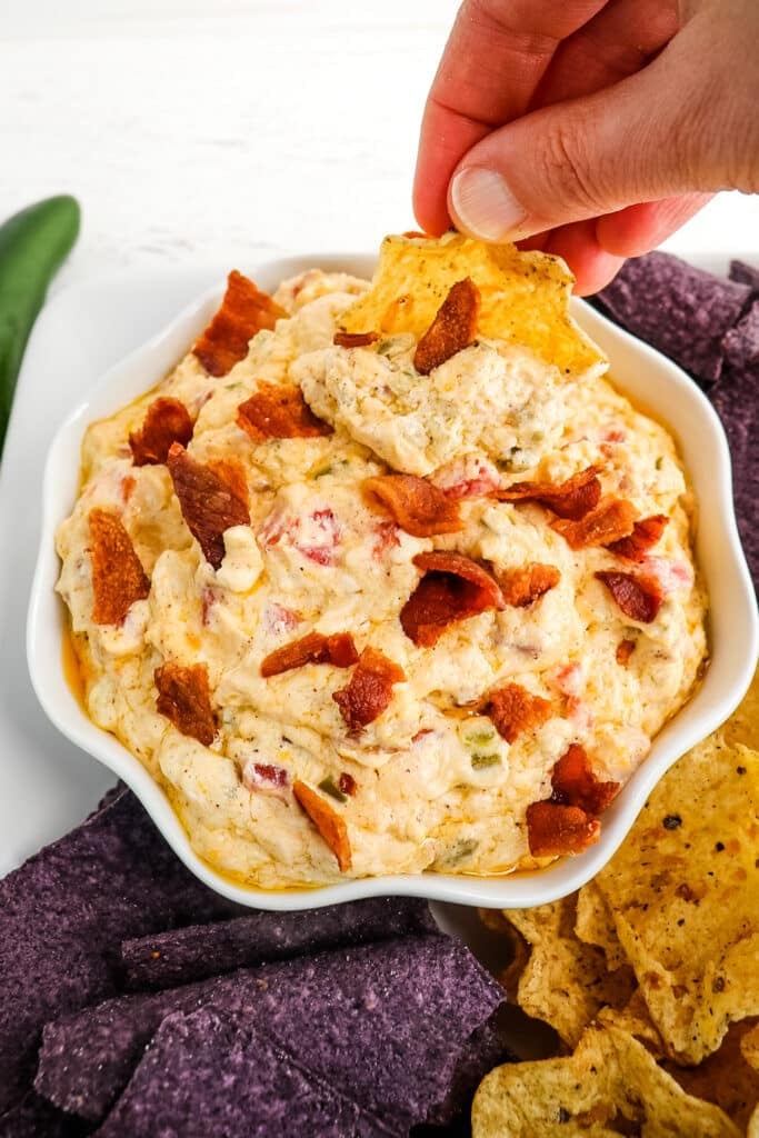 Jalapeno cream cheese dip, in a bowl, topped with bacon and a tortilla chip scooping out a serving.