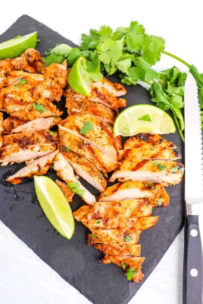 Sliced pollo asado on cutting board with knife, lime wedges and cilantro on the side.