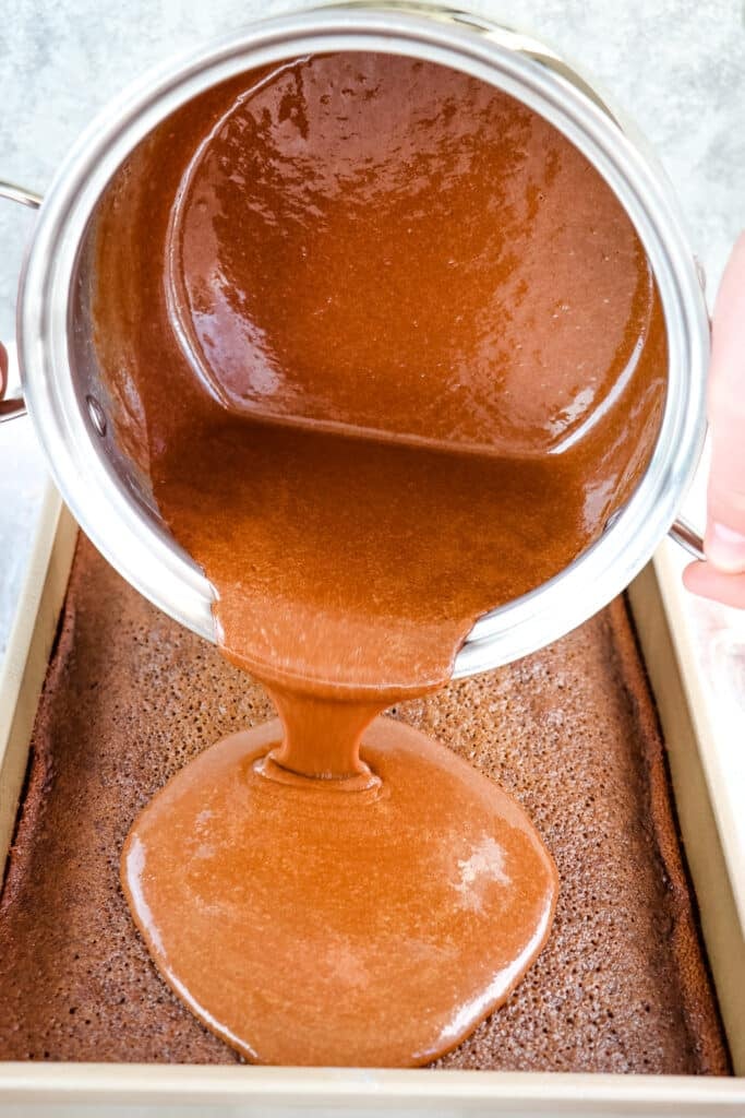Chocolate frosting being poured from saucepan onto baked chocolate cake with buttermilk.