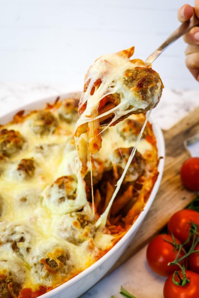 Serving of meatball casserole being taken with spoon from baking dish, with cheese pulls.