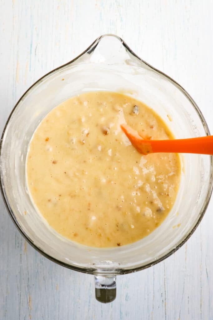 Mixed banana bread batter in a mixing bowl with a rubber scraper in the bowl.