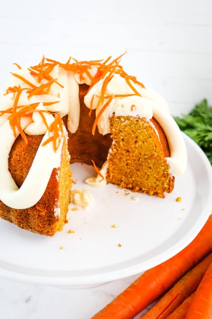 Carrot cake budnt cake with slices removed on a cake plate, topped with cream cheese frosting and carrot strands.