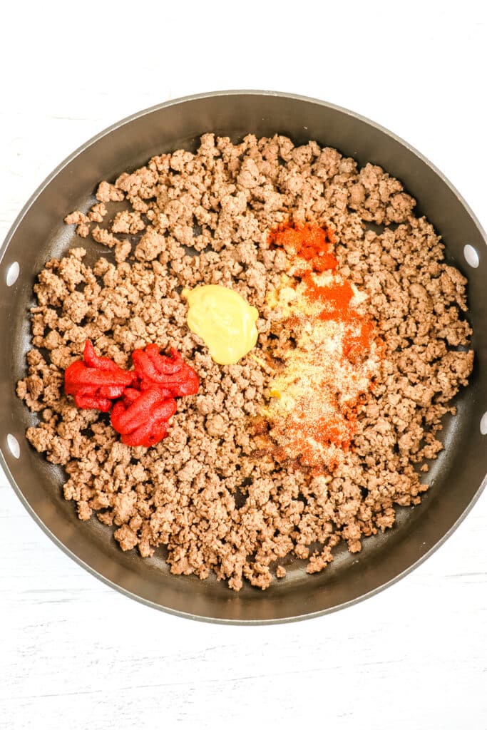 Browned ground beef, tomato paste, mustard and spices in skillet for cheeseburger macaroni.